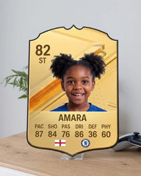 Personalised FC27 Gold football card standing on a clear acrylic stand on a wooden desk next to a game controller